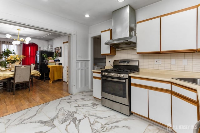 kitchen featuring light countertops, stainless steel range with gas stovetop, a notable chandelier, white cabinetry, and wall chimney range hood