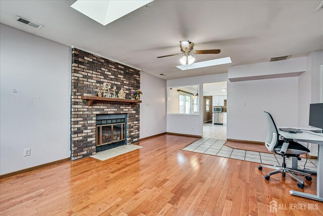 unfurnished office featuring a skylight, wood-type flooring, baseboards, a brick fireplace, and a ceiling fan