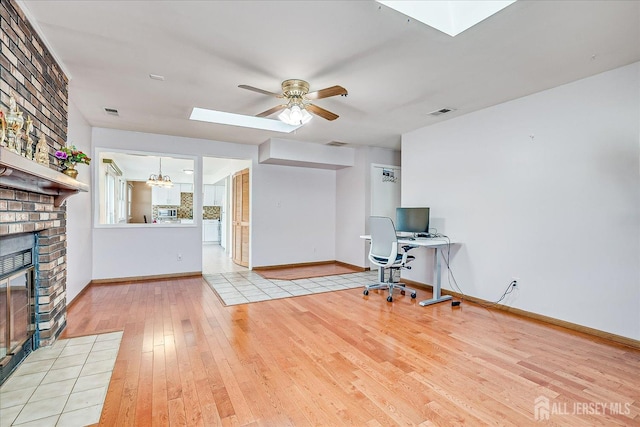unfurnished office featuring a skylight, wood-type flooring, ceiling fan, a brick fireplace, and baseboards