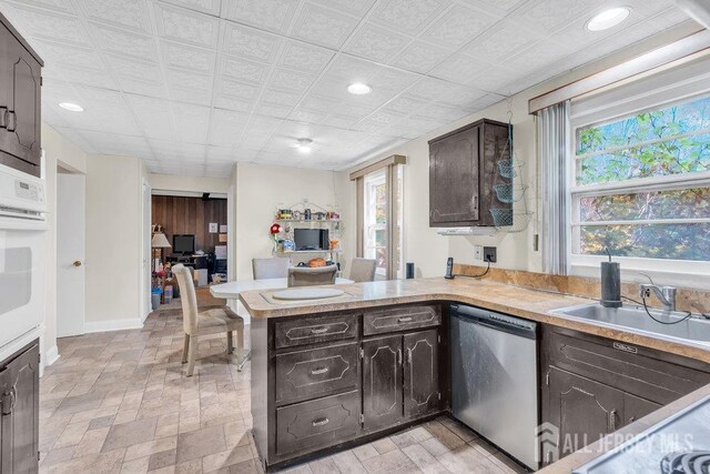 kitchen featuring white oven, light countertops, stainless steel dishwasher, dark brown cabinetry, and a peninsula