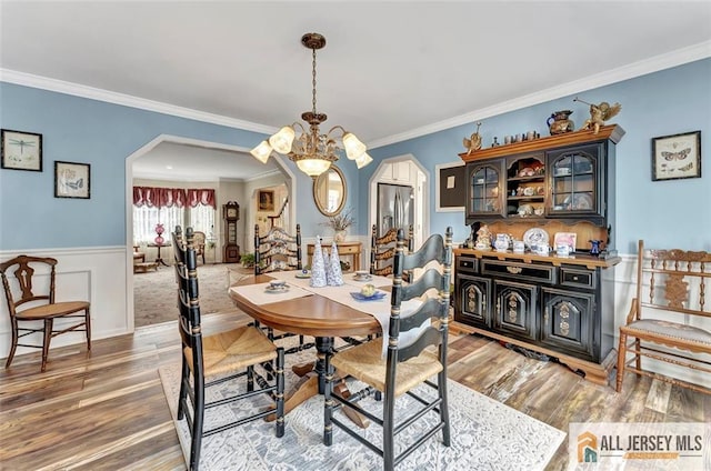 dining room featuring arched walkways, wood finished floors, ornamental molding, a chandelier, and a wainscoted wall