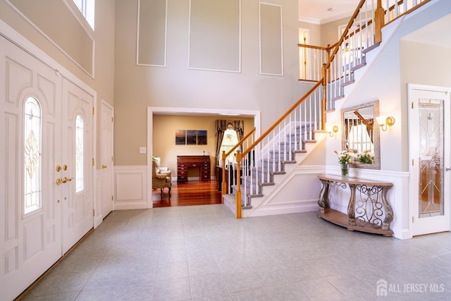 tiled entryway with a towering ceiling, plenty of natural light, stairway, a decorative wall, and wainscoting