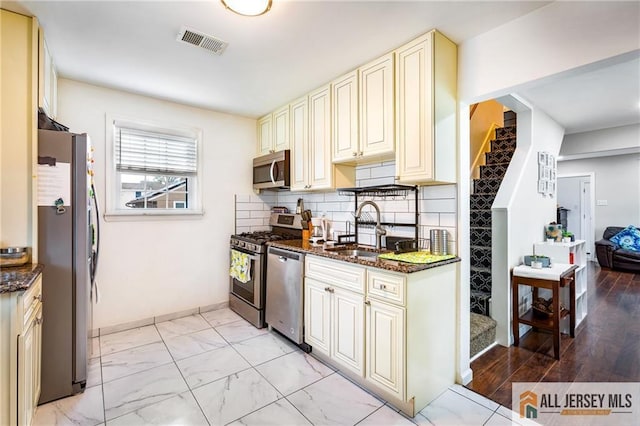 kitchen with a sink, cream cabinetry, stainless steel appliances, and dark stone counters