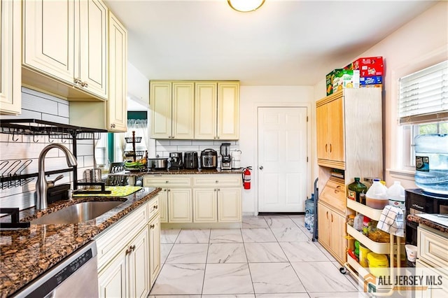 kitchen with light marble finish floors, dishwasher, a sink, cream cabinetry, and dark stone countertops