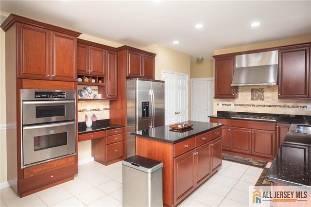 kitchen with wall chimney exhaust hood, a kitchen island, dark stone counters, light tile patterned floors, and appliances with stainless steel finishes