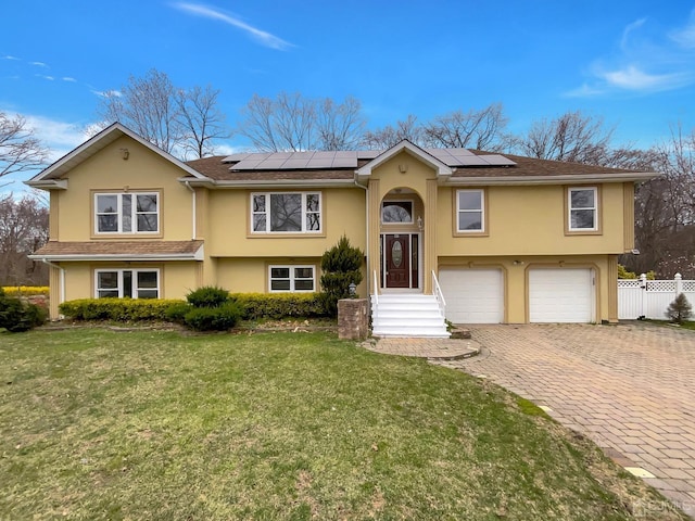 split foyer home featuring a front lawn, fence, roof mounted solar panels, stucco siding, and decorative driveway