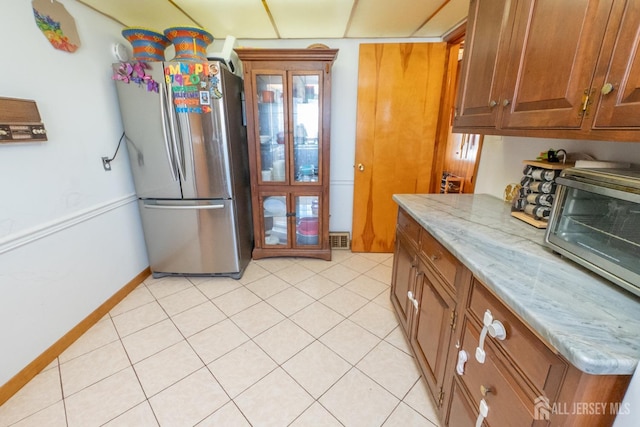 kitchen featuring light tile patterned floors, visible vents, brown cabinetry, light stone counters, and freestanding refrigerator