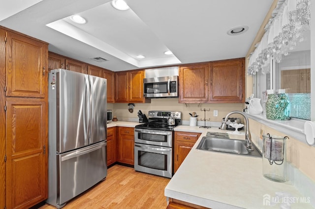 kitchen with stainless steel appliances, a sink, brown cabinetry, a raised ceiling, and recessed lighting