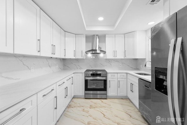 kitchen with sink, appliances with stainless steel finishes, white cabinetry, a tray ceiling, and wall chimney exhaust hood