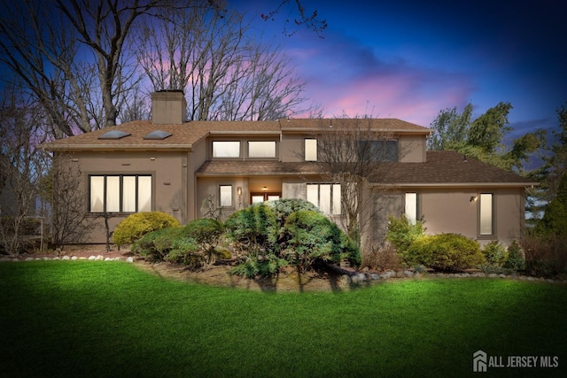 back of property at dusk with a chimney, stucco siding, and a lawn