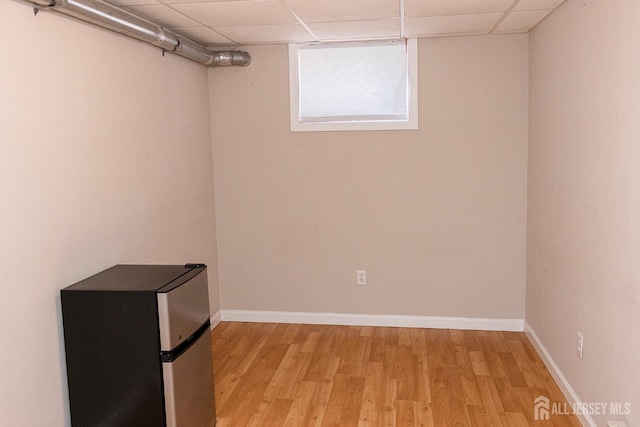 below grade area with freestanding refrigerator, light wood-style floors, baseboards, and a drop ceiling