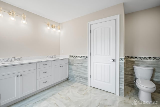 full bath with a wainscoted wall, toilet, marble finish floor, tile walls, and a sink