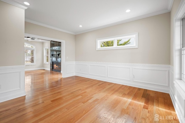 empty room featuring a wainscoted wall, ornamental molding, a glass covered fireplace, recessed lighting, and light wood-style floors