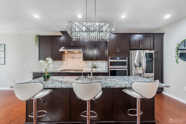 kitchen with dark wood-type flooring, appliances with stainless steel finishes, hanging light fixtures, backsplash, and an island with sink