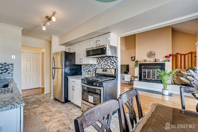 kitchen featuring ornamental molding, white cabinets, appliances with stainless steel finishes, a glass covered fireplace, and tasteful backsplash