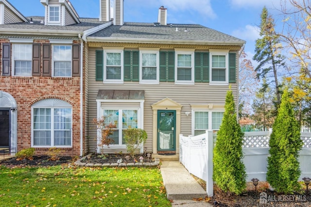 multi unit property with roof with shingles, brick siding, a chimney, and fence