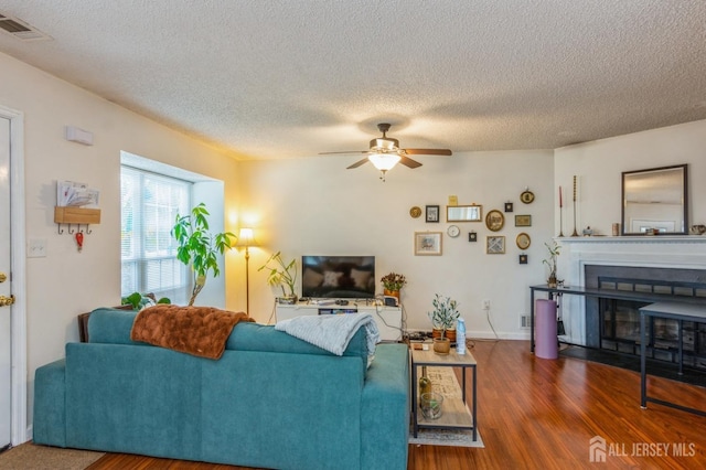 living area featuring ceiling fan, a textured ceiling, a fireplace with flush hearth, wood finished floors, and visible vents