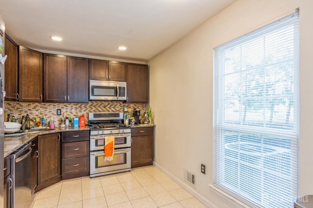 kitchen featuring dark brown cabinetry, stainless steel appliances, a sink, light stone countertops, and tasteful backsplash