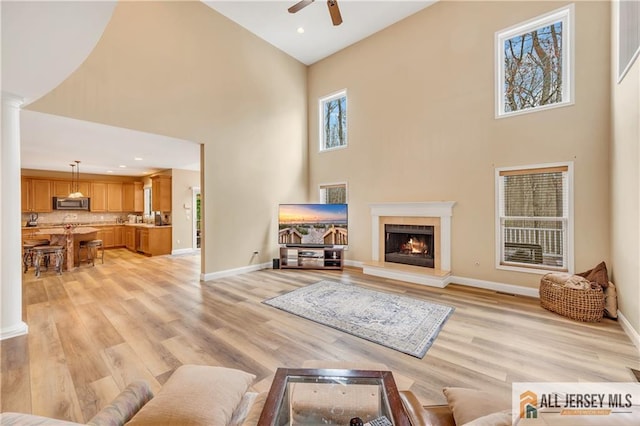 living room with a towering ceiling, a ceiling fan, light wood-style flooring, decorative columns, and baseboards