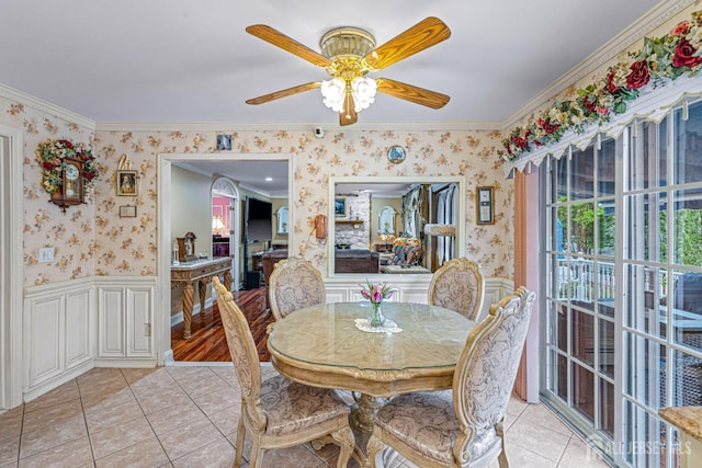 dining space with wallpapered walls, crown molding, light tile patterned flooring, ceiling fan, and a wainscoted wall