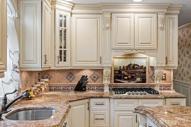 kitchen featuring stainless steel gas stovetop, cream cabinetry, a sink, glass insert cabinets, and light stone countertops