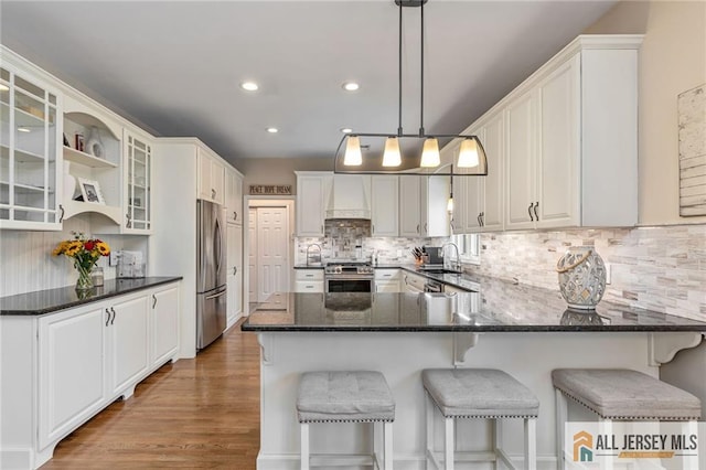 kitchen with stainless steel appliances, a peninsula, tasteful backsplash, wall chimney exhaust hood, and glass insert cabinets