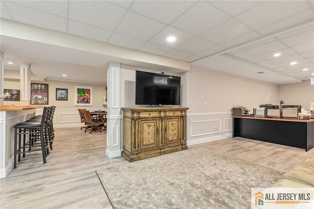living room with light wood finished floors, a decorative wall, a paneled ceiling, wainscoting, and a bar