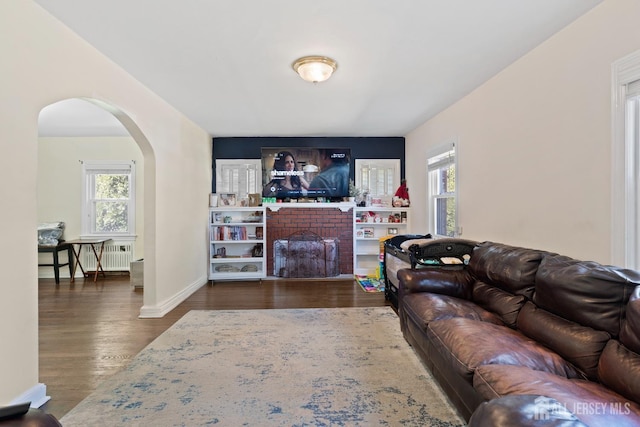 living room featuring radiator heating unit, wood finished floors, arched walkways, baseboards, and a brick fireplace