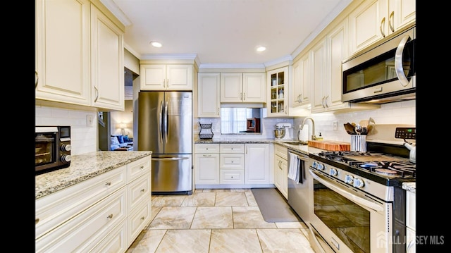 kitchen with a sink, light stone counters, backsplash, appliances with stainless steel finishes, and glass insert cabinets