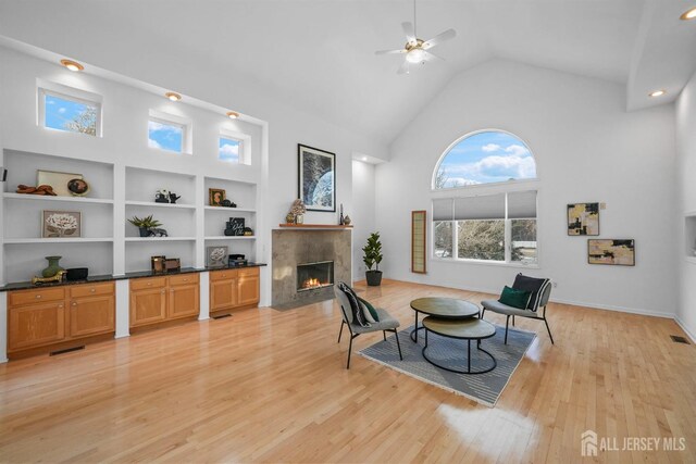 living room featuring high vaulted ceiling, light wood-style flooring, a ceiling fan, a fireplace with flush hearth, and visible vents