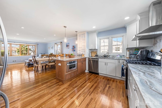 kitchen featuring a peninsula, wall chimney range hood, stainless steel appliances, light wood finished floors, and plenty of natural light