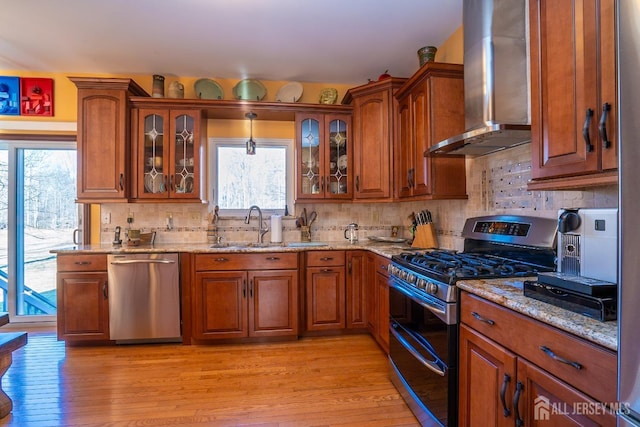 kitchen with sink, hanging light fixtures, stainless steel appliances, wall chimney range hood, and light wood-type flooring