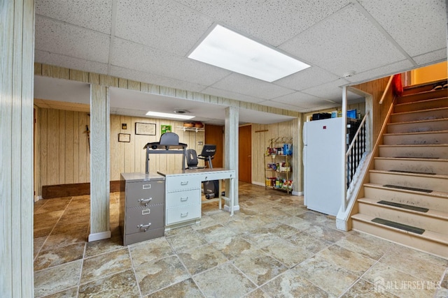 basement featuring wooden walls, a drop ceiling, and white fridge