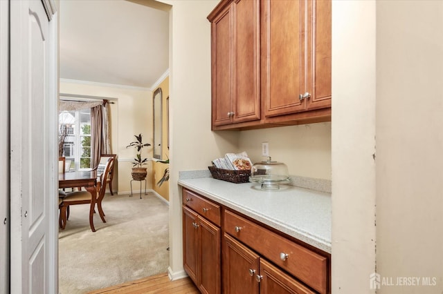 kitchen with light carpet, baseboards, brown cabinetry, ornamental molding, and light countertops