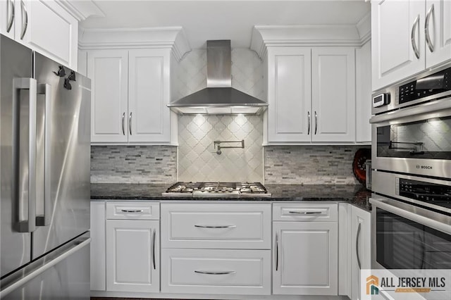 kitchen with appliances with stainless steel finishes, wall chimney range hood, white cabinets, and backsplash