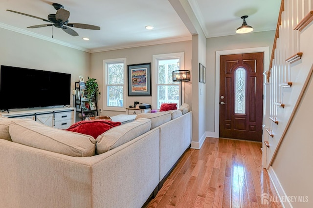 living area featuring light wood-style floors, baseboards, crown molding, a ceiling fan, and stairs