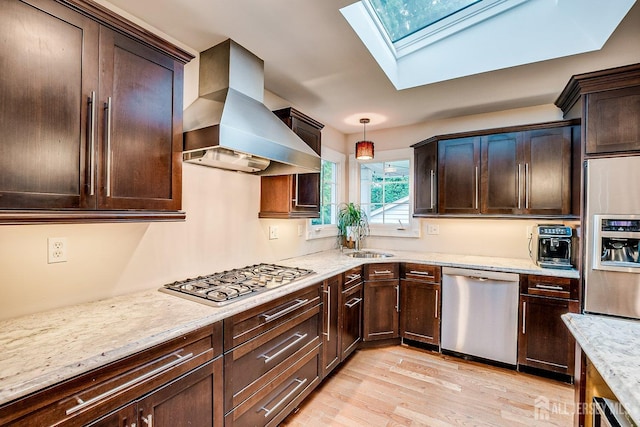kitchen with stainless steel appliances, wall chimney range hood, a skylight, light wood-style floors, and light stone countertops