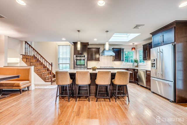 kitchen featuring appliances with stainless steel finishes, wall chimney range hood, light countertops, light wood-style flooring, and a breakfast bar area