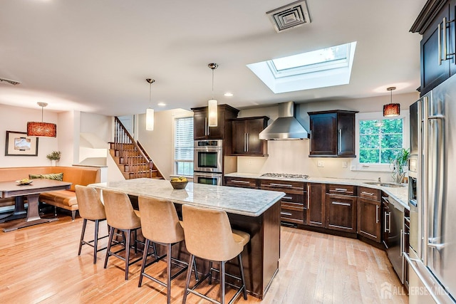 kitchen with wall chimney range hood, stainless steel appliances, a center island, a kitchen breakfast bar, and light wood-style flooring