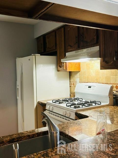 kitchen featuring under cabinet range hood, dark stone counters, white appliances, decorative backsplash, and dark brown cabinets