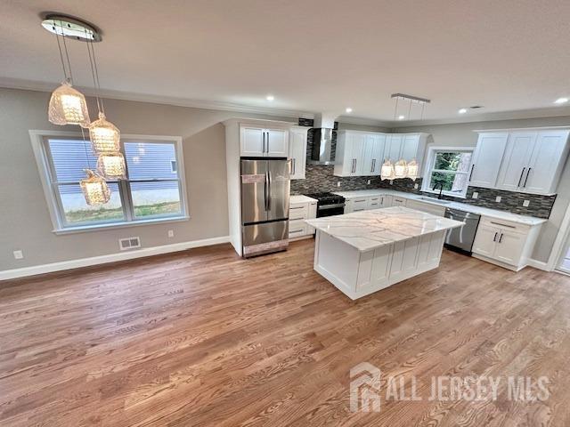 kitchen featuring a kitchen island, white cabinets, pendant lighting, stainless steel appliances, and wall chimney range hood