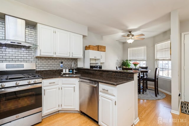 kitchen with a sink, white cabinetry, appliances with stainless steel finishes, wall chimney range hood, and ceiling fan