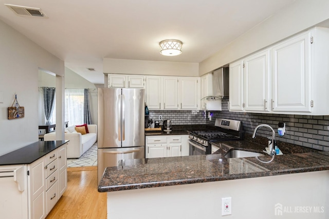 kitchen with light wood finished floors, dark stone counters, stainless steel appliances, wall chimney exhaust hood, and a sink