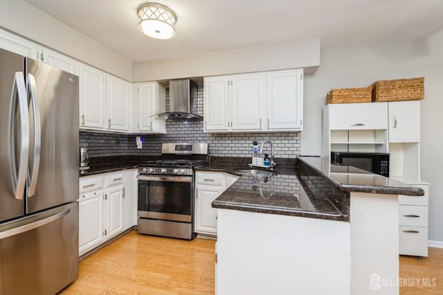 kitchen with a peninsula, a sink, light wood-style floors, appliances with stainless steel finishes, and wall chimney exhaust hood