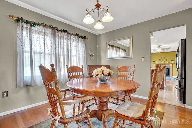 dining area with baseboards, a fireplace with raised hearth, ceiling fan with notable chandelier, and light wood-style floors