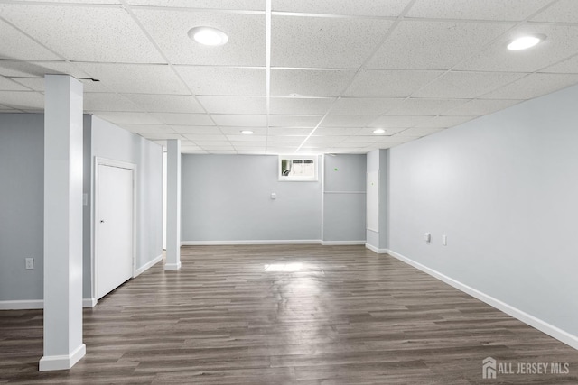 below grade area with an AC wall unit, a drop ceiling, dark wood finished floors, and baseboards