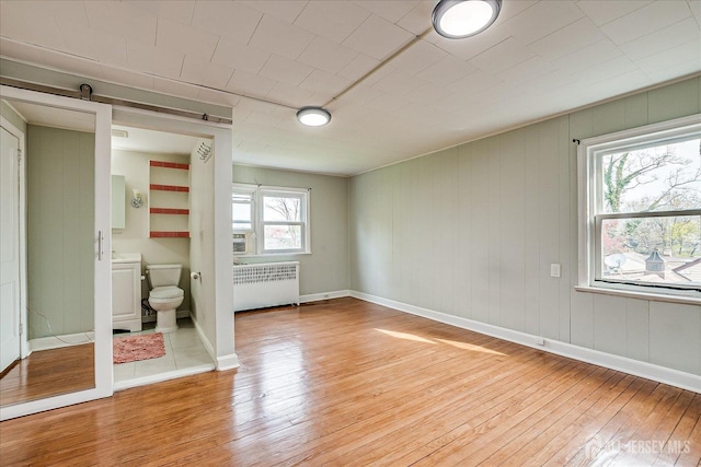 unfurnished bedroom featuring radiator, light wood finished floors, a barn door, baseboards, and ensuite bathroom