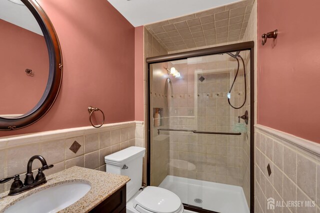 bathroom with toilet, vanity, wainscoting, a stall shower, and tile walls