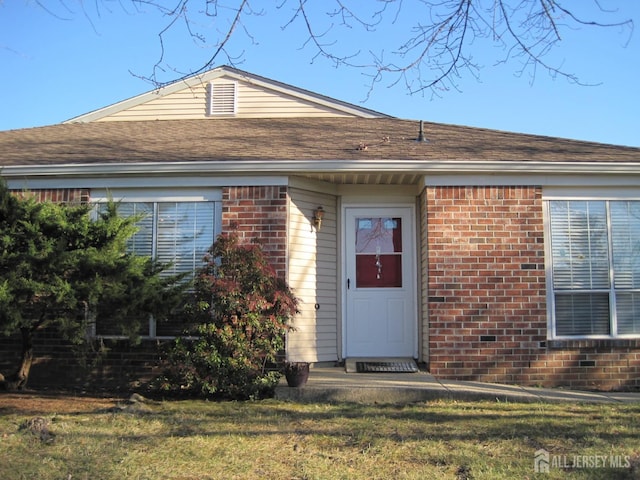 view of exterior entry with a yard, a shingled roof, and brick siding