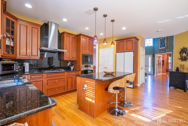 kitchen featuring light wood-type flooring, a kitchen island, pendant lighting, stainless steel appliances, and wall chimney range hood